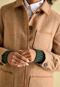 Brown houndstooth coat with a collar, button closure, and chest pocket. Features contrasting tan trim and green ribbed cuffs.