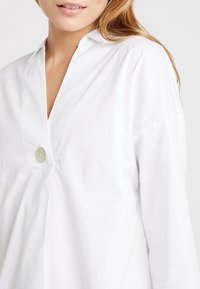 Weiße Baumwollbluse mit V-Ausschnitt, langen Ärmeln und einem dekorativen Knopf auf der Vorderseite. Glatte Textur, minimalistisches Design.