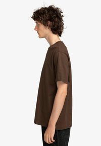Camiseta de algodón marrón con mangas cortas, escote redondo y corte holgado. Modelo mostrado de perfil, vistiendo pantalones oscuros. Diseño simple, sin patrones visibles.