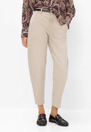 Pantaloni beige a vita alta con gamba affusolata e tasche frontali, abbinati a una camicetta paisley, cintura nera e mocassini in pelle neri.