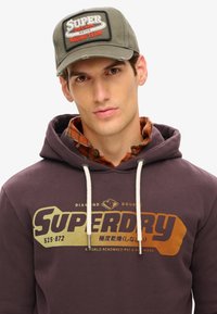 Fioletowy sweatshirt z grafiką "Superdry", białymi sznurkami do ściągania, nałożony na koszulę w kratę z guzika. Szara czapka z logo "Racing Team".
