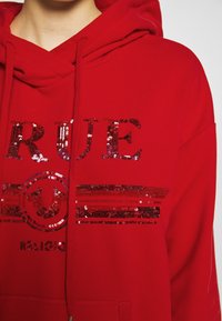 Červená mikina s kapucí s flitrovým nápisem "RUE" a kruhovým logem. Obsahuje stahovací kapuci a přední kapsu. Měkká, strukturovaná tkanina.