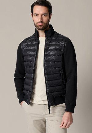 Mężczyzna z krótkimi ciemnymi włosami i brodą nosi czarną pikowaną kurtkę na beżowej koszuli i beżowych spodniach, stoi na jednolitym tle.