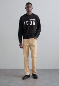 Sweatshirt noir avec le texte blanc "ICON", pantalon beige et chaussures noires ; le modèle se tient devant un fond neutre.
