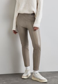 Kampanj av taupe-faux läderbyxor med sömsömnad, slim fit och elastisk midja. Kombinerat med vita och beige sneakers på ett strukturerat golv.