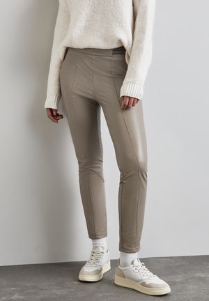 Pantaloni in finta pelle color taupe con dettagli sulle cuciture, fit slim e vita elastica. Abbinati a sneaker bianche e beige su un pavimento testurizzato.