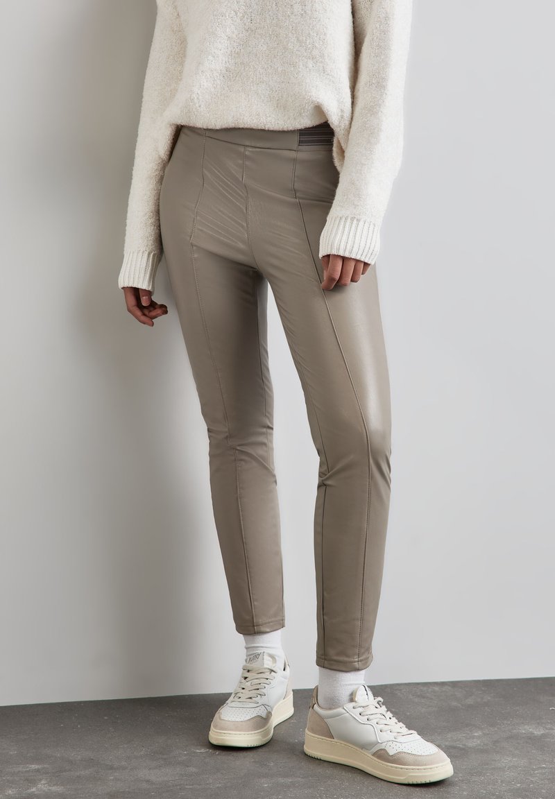 Kampanj av taupe-faux läderbyxor med sömsömnad, slim fit och elastisk midja. Kombinerat med vita och beige sneakers på ett strukturerat golv.