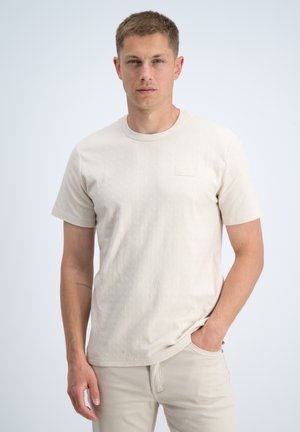 Jonge man staat met één hand in zijn zak, draagt een licht beige gestructureerd kort mouwen overhemd en bijpassende broek tegen een effen achtergrond.