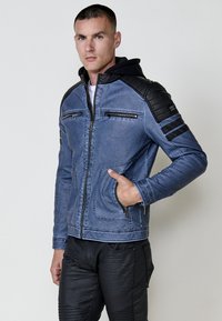 Uomo che indossa una giacca blu con zip, caratterizzata da toppe trapuntate nere sulle spalle e sulle maniche, in piedi con una mano in tasca su uno sfondo neutro.