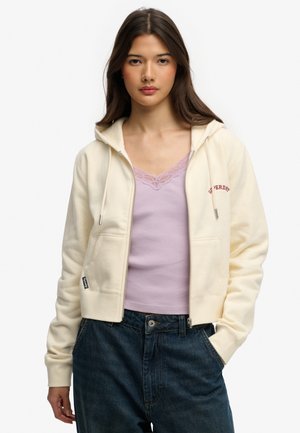 Jonge vrouw met lang bruin haar, een crèmekleurige hoodie met ritssluiting over een lila top met kanten rand en donkerblauwe spijkerbroek, staand met één hand in de zak.