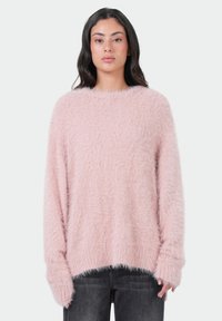 Vrouw met lang donker haar, gekleed in een oversized lichtroze fuzzy trui en donkergrijze jeans, staande tegen een effen witte achtergrond.