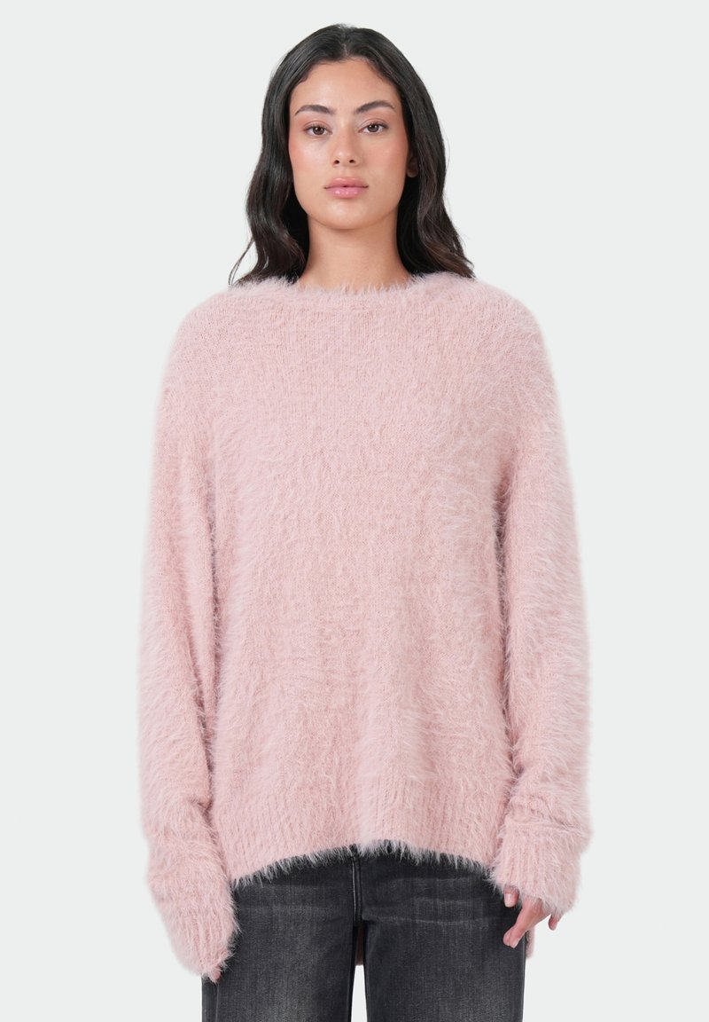 Vrouw met lang donker haar, gekleed in een oversized lichtroze fuzzy trui en donkergrijze jeans, staande tegen een effen witte achtergrond.