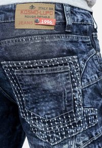 Jeans in denim di un blu indaco scuro con cuciture bianche a contrasto, dotati di tasche a toppa e un'etichetta in pelle con marchio sulla vita.