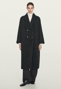 Manteau noir à double boutonnage en tissu texturé, avec des revers crantés, des manches surdimensionnées et deux boutons à l'avant. Porté sur un top blanc.