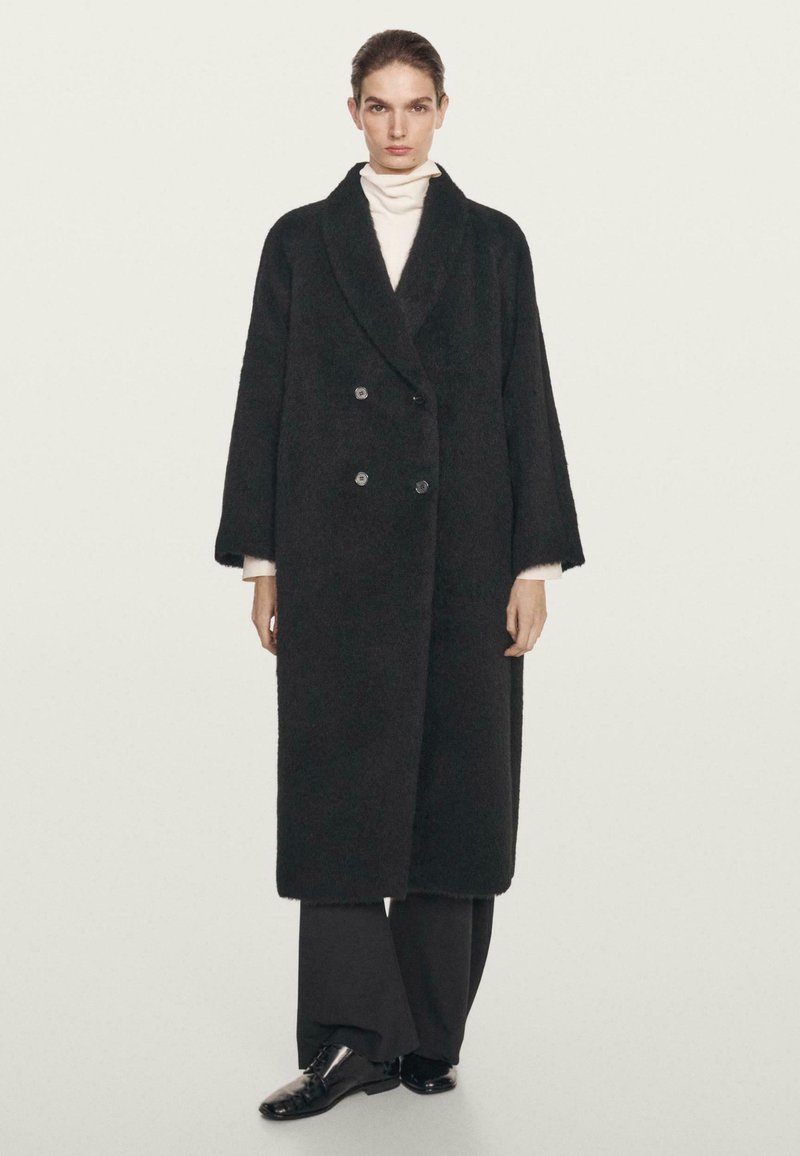 Manteau noir à double boutonnage en tissu texturé, avec des revers crantés, des manches surdimensionnées et deux boutons à l'avant. Porté sur un top blanc.