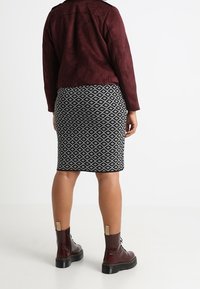 Giacca bordeaux in finto suede, gonna con motivo geometrico bianco e nero, e stivaletti marroni con dettagli dorati. Vista posteriore che mostra l'outfit.