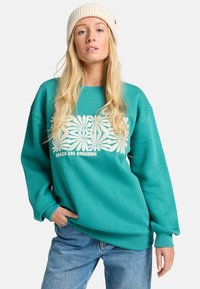 Felpa turchese con design grafico floreale bianco e testo "RAGGIUNGI IL SOLE." Polsini e colletto a costine, abbinata a jeans azzurri.