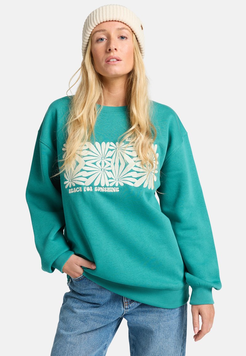 Felpa turchese con design grafico floreale bianco e testo "RAGGIUNGI IL SOLE." Polsini e colletto a costine, abbinata a jeans azzurri.