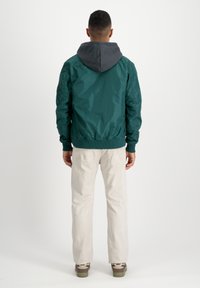 Groene bomberjack met grijze capuchon, elastische manchetten en zoom. Gelaagd met lichtgekleurde broek en schoenen met groene zolen, wat een casual uitstraling geeft.