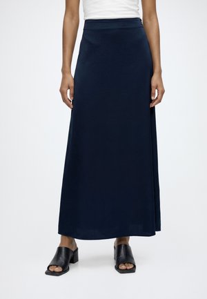 Femme portant une longue jupe bleu marine, un haut blanc sans manches et des chaussures noires à talons ouvertes, debout devant un fond clair uni.