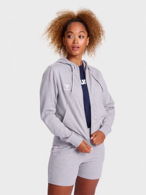 Femme en train de dézipper un sweat à capuche gris clair, portant un short assorti et un t-shirt bleu marine, debout devant un fond clair uni.