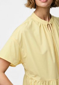 Vestido amarillo de manga corta con cuello alto, que presenta detalles fruncidos en el cuello y la cintura, hecho de una tela suave y ligera.