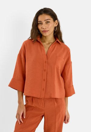 Mujer con cabello largo y oscuro, vistiendo una camisa abotonada color óxido naranja con mangas 3/4 y pantalones a juego, de pie frente a un fondo blanco.