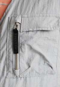 Gilet grigio chiaro con una tasca prominente con cerniera dotata di finiture argentate, tessuto strutturato e tirante nero a contrasto.