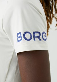 Kreem värvi lühikeste varrukatega särk sinise täpikujundusega "BORG" vasakul varrukal. Sile kangas, istuv disain. Ei mingeid täiendavaid mustreid ega aktsente.