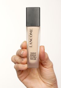 Flacon de fond de teint Lancôme avec un corps en verre transparent rectangulaire et un capuchon carré noir. L'étiquette indique "Teint Idole Ultra Wear" en lettres noires épaisses.