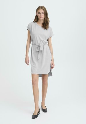 Noor noor seisab seljas heleda halli vööga lühikeste varrukatega kleidi ja mustade madalate kontsadega kingadega, puhta heleda taustaga.