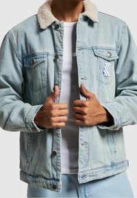 Persona vistiendo una chaqueta de denim azul claro con cuello de borreguito y camiseta blanca de canalé, sosteniendo los bordes de la chaqueta con ambas manos.