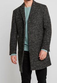 Manteau en laine grise texturée avec un col en V et une poche avant unique, superposé à un sweat-shirt teal, porté avec un pantalon noir.