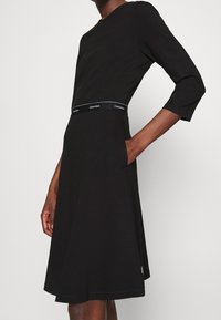 Černé šaty s přiléhavým vrškem, tříčtvrtečními rukávy, rozšířenou sukní a páskem s logem Calvin Klein. Měkká látka s hladkým povrchem.