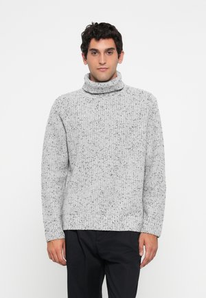 Uomo che indossa un maglione a collo alto grigio chiaro a pois e pantaloni neri, in piedi davanti a uno sfondo bianco semplice.
