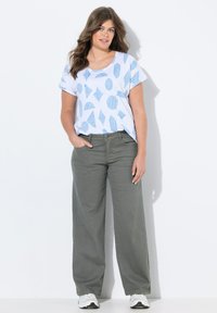 T-shirt blanc avec des motifs géométriques bleus, associé à un pantalon ample vert olive. Le modèle porte des baskets blanches. L'arrière-plan est blanc uni.