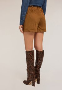 Bruine suède shorts met opgerolde mouwen, gecombineerd met kniehoge boots met een snakeskin print en een blokhak. Een denim overhemd is gedeeltelijk zichtbaar.