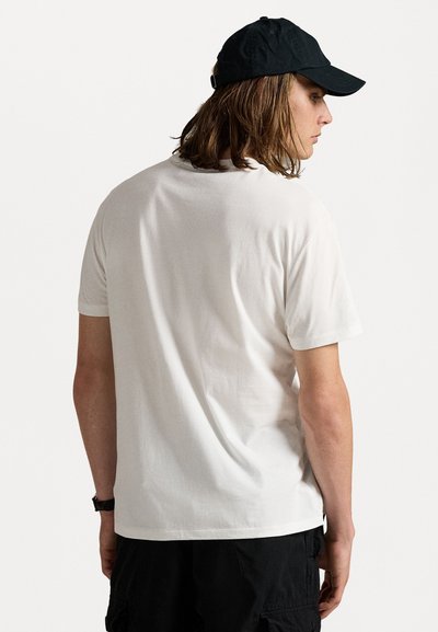 Tricou alb din bumbac, cu mâneci scurte, textură fină și guler în formă de cerc. Asortat cu o șapcă neagră, văzut din spate. Stil și tăietură casual.