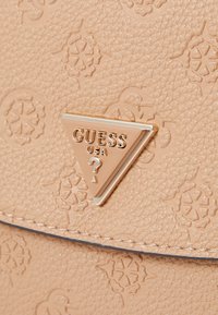 Svijetlosmeđi teksturirani kožni materijal s reljefnim cvjetnim uzorcima, s trokutastom zlatnom oznakom na kojoj piše "GUESS USA."