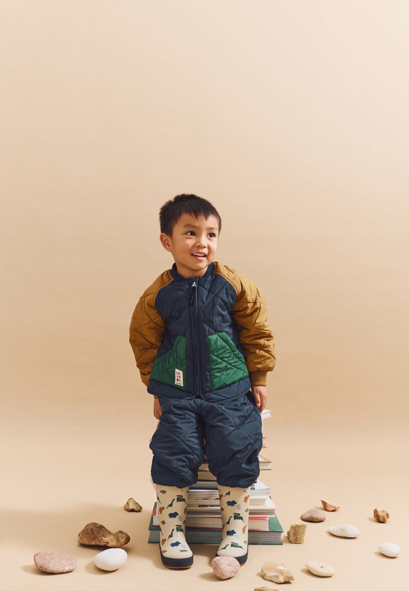 Quilted jacket with green and brown sections, blue pants, and dinosaur-patterned rain boots. Child stands on stacked books.