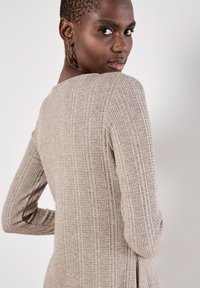 Vestido de punto de manga larga con canalé en color beige claro, con un escote redondeado. Presenta una silueta ajustada y una textura suave; se lleva con pendientes de aro.