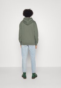 Sudadera verde con capucha de corte relajado, llevada sobre unos vaqueros de lavado claro. La persona lleva botas verdes hasta el tobillo, con un fondo de tela texturada.