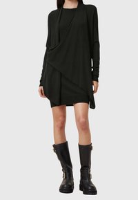 Femme portant une robe noire à manches longues avec un tissu drapé en couches et des bottes en cuir noires montant jusqu'aux genoux, ornées de boucles.