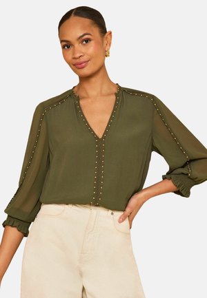 Žena v olivovo zelenej blúzke so zlatými nitmi pozdĺž výstrihu a rukávov, v kombinácii s béžovými nohavicami s vysokým pásom.