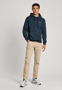 Felpa con cappuccio navy, dotata di tasca frontale e lacci, abbinata a pantaloni beige e sneakers grigie. Presenta un branding minimale sul petto.