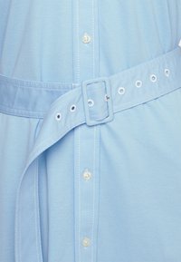Ljusblå skjorta med knäppning framtill, ett tygbälte med rektangulär spänne och kontrasterande vita sömmar.