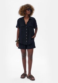 Czarna koszula z krótkim rękawem, z zapięciem na guziki oraz dwiema przednimi kieszeniami, zestawiona z pasującymi szortami. Model nosi brązowe sandały na platformie.