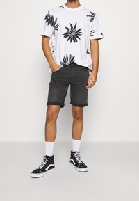 Hombre con camiseta blanca con grandes estampados florales negros, pantalones cortos de mezclilla negros arremangados, calcetines blancos y zapatillas altas negras.