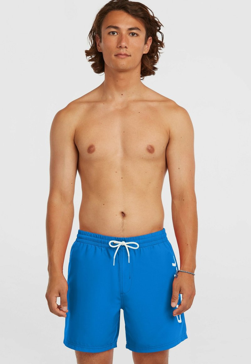 Muž bez trička se středně dlouhými hnědými vlasy, oblečený v modrých plavkových šortkách s bílým šňůrkem, stojí před jednobarevným pozadím.