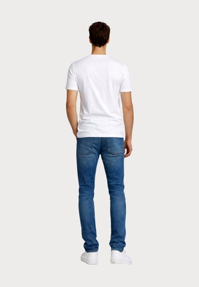 Άνδρας που φοράει απλό λευκό κοντομάνικο μπλουζάκι, μπλε τζιν και λευκά αθλητικά παπούτσια, στέκεται με την πλάτη προς το φακό μπροστά σε ένα απλό φόντο.